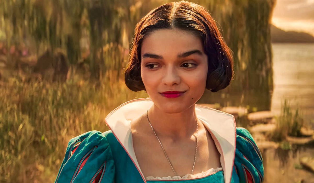 Une femme vêtue d'une robe sarcelle aux manches bouffantes se tient à l'extérieur, près de l'eau et de la verdure, regardant sur le côté avec un léger sourire, rappelant la gracieuse interprétation de Rachel Zegler dans Blanche-neige.