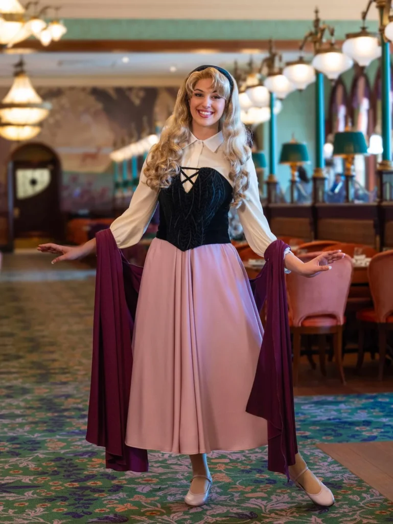 Une femme habillée en princesse de conte de fées avec une longue jupe rose, un chemisier crème et un corset noir sourit dans la salle à manger de style ancien du restaurant Regal View à Disneyland Paris, célébrant les nouvelles tenues Ariel Mulan Aurore.
