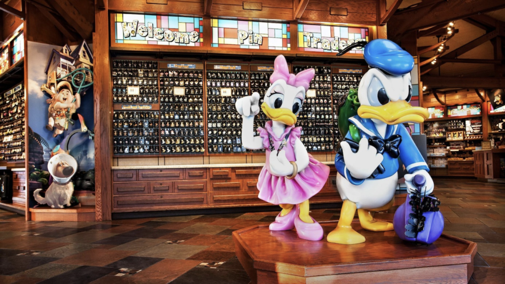 Un présentoir avec de grandes statues de Daisy Duck et Donald Duck se trouve devant un mur rempli de pin's à collectionner, célébrant le Pin Trading à Disneyland Paris à l'intérieur d'une boutique très éclairée.