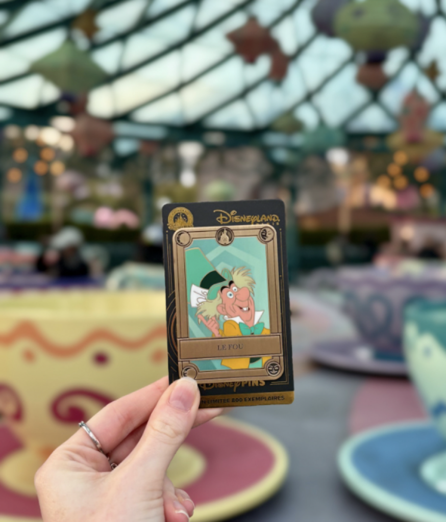 Une main tient une carte à collectionner de Disneyland Paris représentant le Chapelier fou, clin d'œil à Pin Trading, avec les attractions du manège à thé floues en arrière-plan, capturant un moment magique du 6 octobre 2001.
