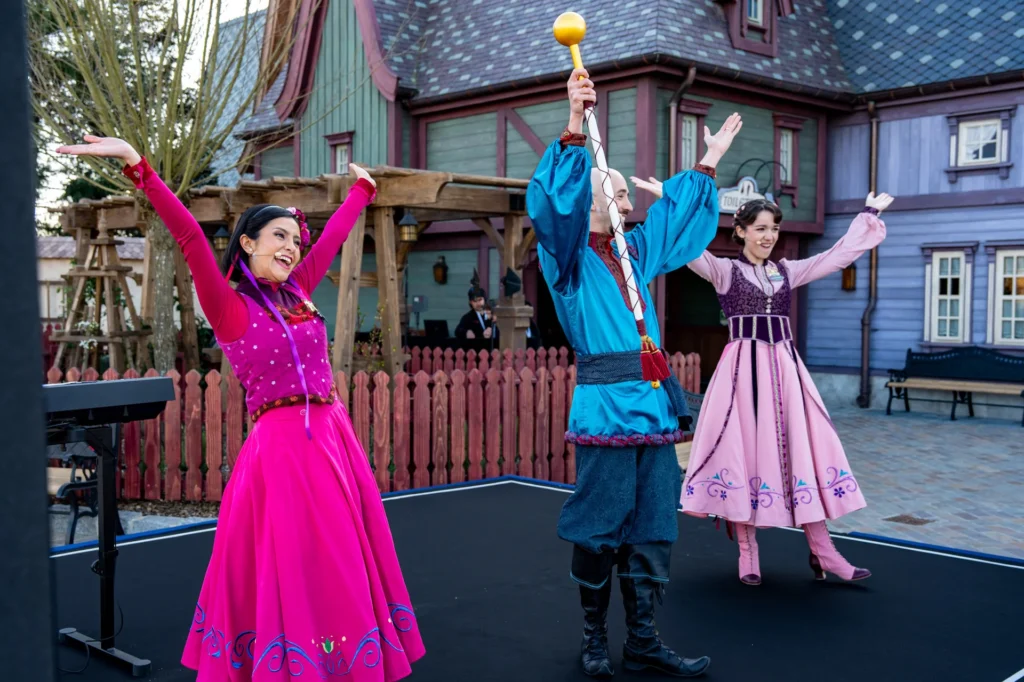 Trois artistes en costumes colorés se tiennent sur une scène extérieure, les bras levés, souriant devant un bâtiment aux murs violets et verts inspirés d'Arendelle, célébrant la magie de World Of Frozen à Disneyland Paris.