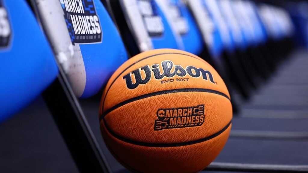 Un ballon de basket Wilson portant le logo de la March Madness repose sur une rangée de chaises bleues étiquetées "March Madness" dans une arène sportive, illustrant l'excitation de NCAA 2026 et l'esprit du basket-ball américain.
