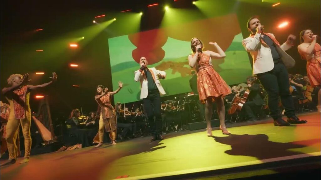 Un groupe de chanteurs se produit sur scène avec un orchestre en arrière-plan, des éclairages colorés et un grand écran affichant des images abstraites pendant le ciné-concert symphonique de Disney En Concert.