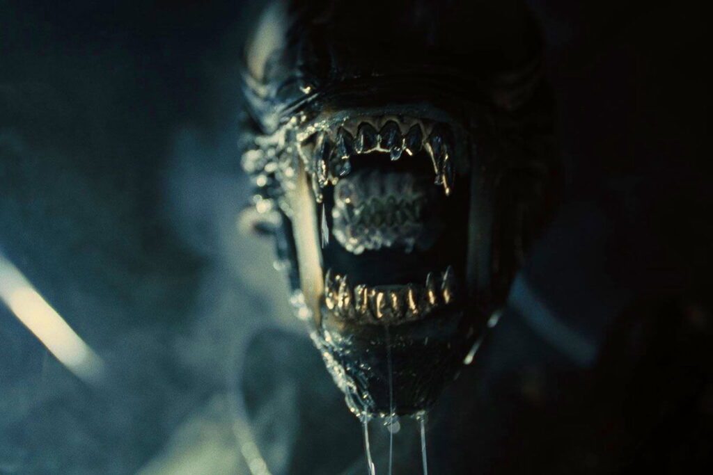 Gros plan d'une créature à la bouche métallique et dentée grande ouverte, montrant des dents pointues et des gouttes de salive, sur un fond sombre - évoquant l'atmosphère glaçante d'Alien Romulus 2 par la suite de Michael Sarnoski.