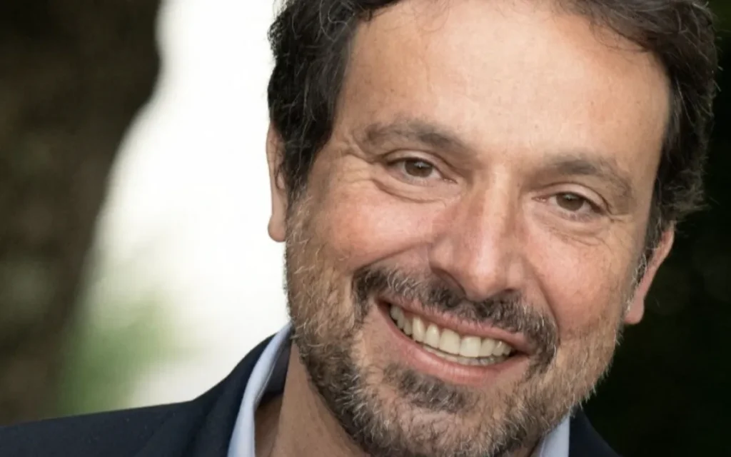 Un homme aux cheveux noirs et à la barbe sourit à la caméra, vêtu d'une veste sombre et d'une chemise claire, sur un fond extérieur flou. Il s'agit de Bruno Salomone, connu pour ses rôles de voix chez Disney et Doublages Pixar.