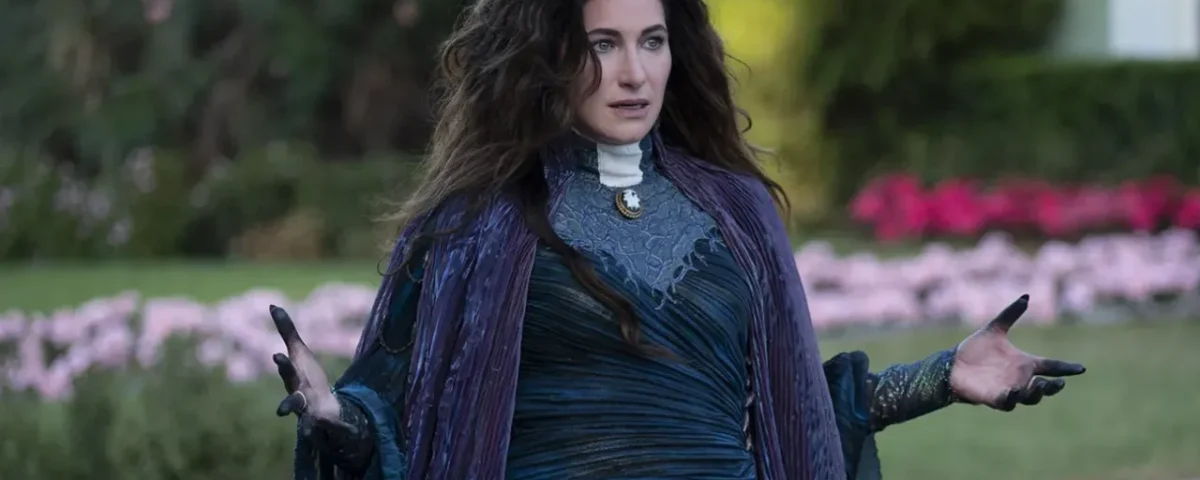 Une femme aux longs cheveux noirs, rappelant l'interprétation de Kathryn Hahn en Mère Gothel dans le remake de Raiponce, se tient à l'extérieur dans une tenue fluide violet foncé et bleu, les mains légèrement levées, avec de la verdure et des fleurs roses floues derrière elle.