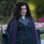 Une femme aux longs cheveux noirs, rappelant l'interprétation de Kathryn Hahn en Mère Gothel dans le remake de Raiponce, se tient à l'extérieur dans une tenue fluide violet foncé et bleu, les mains légèrement levées, avec de la verdure et des fleurs roses floues derrière elle.