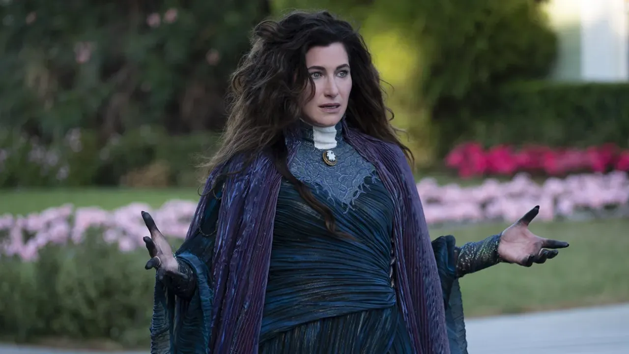 Une femme aux longs cheveux noirs, rappelant l'interprétation de Kathryn Hahn en Mère Gothel dans le remake de Raiponce, se tient à l'extérieur dans une tenue fluide violet foncé et bleu, les mains légèrement levées, avec de la verdure et des fleurs roses floues derrière elle.