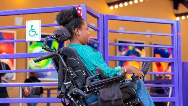 Une femme en fauteuil roulant motorisé se déplace sur une rampe marquée d'un panneau d'accessibilité aux fauteuils roulants, mettant en évidence les aménagements visiteurs à Disneyland Paris, avec des affiches colorées en arrière-plan.