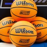 Quatre ballons de basket Wilson EVO NXT avec les logos "March Madness" sont empilés sur un terrain de basket, capturant l'excitation de NCAA 2026 et l'esprit du basket américain pendant la saison des tournois.