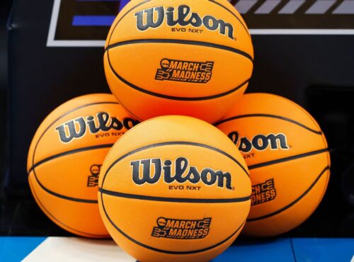 Quatre ballons de basket Wilson EVO NXT avec les logos "March Madness" sont empilés sur un terrain de basket, capturant l'excitation de NCAA 2026 et l'esprit du basket américain pendant la saison des tournois.