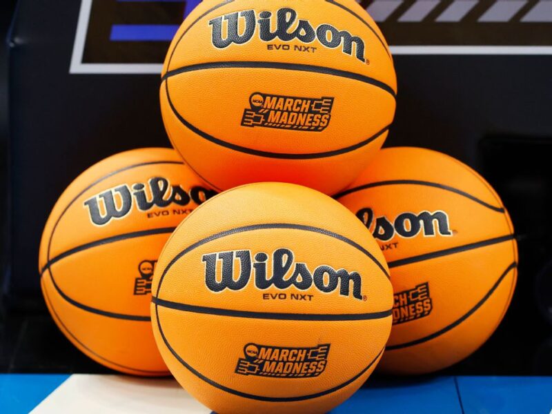 Quatre ballons de basket Wilson EVO NXT avec les logos "March Madness" sont empilés sur un terrain de basket, capturant l'excitation de NCAA 2026 et l'esprit du basket américain pendant la saison des tournois.