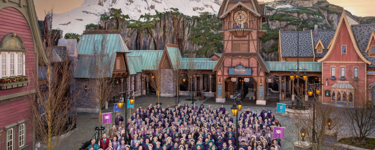 Un grand groupe de personnes en costumes thématiques pose devant une place de village inspirée d'Arendelle, avec des bâtiments alpins et une montagne enneigée en arrière-plan, dans le Monde de Frozen de Disneyland Paris.