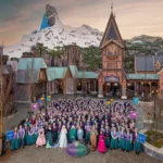 Un grand groupe de personnes en costumes thématiques pose devant une place de village inspirée d'Arendelle, avec des bâtiments alpins et une montagne enneigée en arrière-plan, dans le Monde de Frozen de Disneyland Paris.