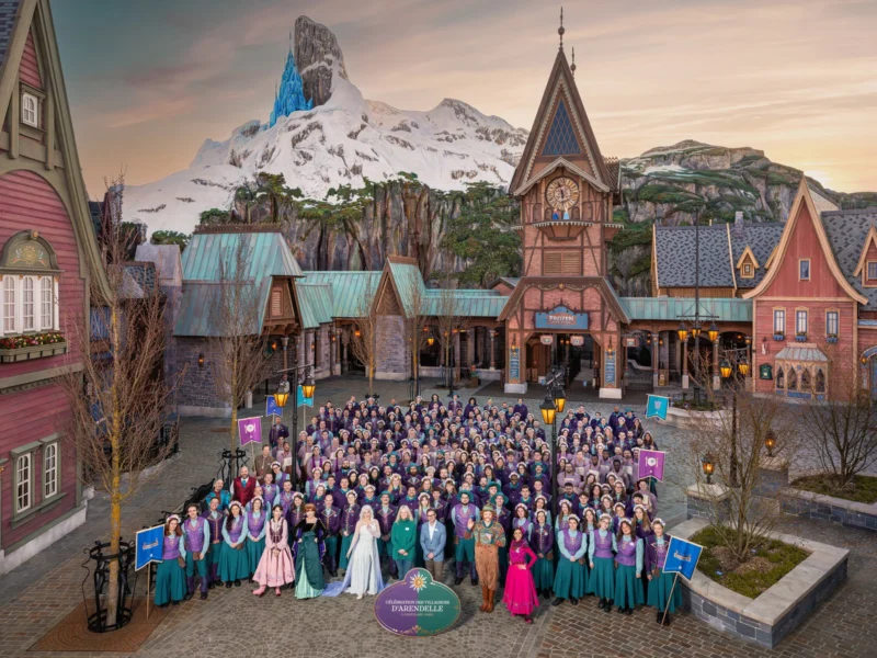 Un grand groupe de personnes en costumes thématiques pose devant une place de village inspirée d'Arendelle, avec des bâtiments alpins et une montagne enneigée en arrière-plan, dans le Monde de Frozen de Disneyland Paris.