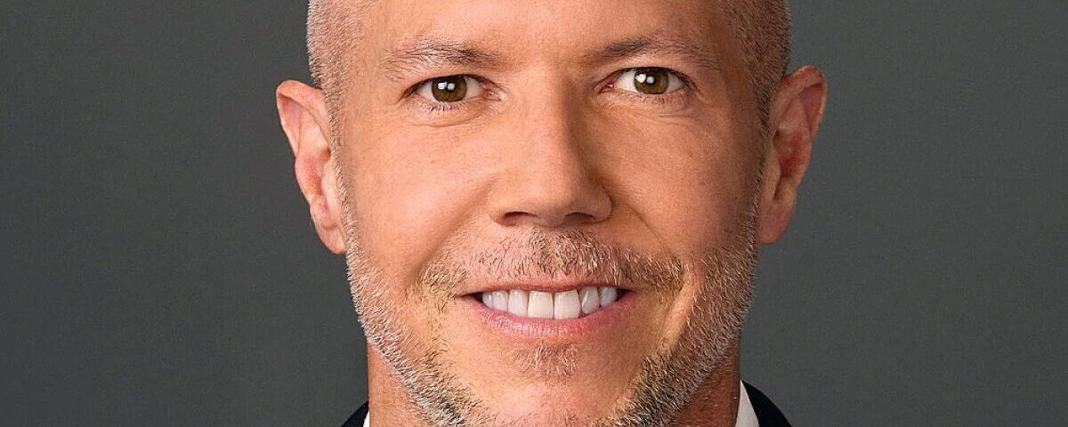 Portrait en gros plan de Paul Roeder, directeur de la communication de Disney, souriant, cheveux courts et clairs, barbe taillée, vêtu d'une veste sombre et d'une chemise blanche, sur un fond gris uni.