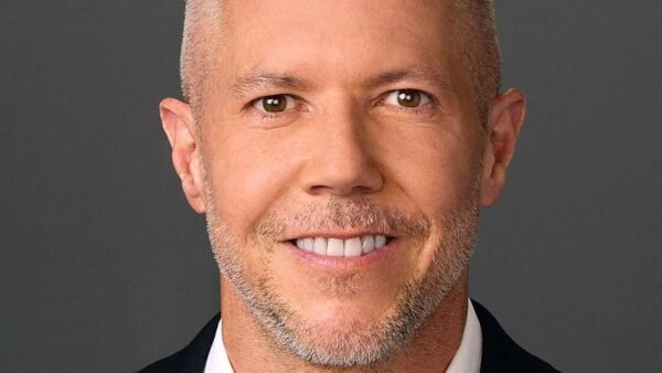 Portrait en gros plan de Paul Roeder, directeur de la communication de Disney, souriant, cheveux courts et clairs, barbe taillée, vêtu d'une veste sombre et d'une chemise blanche, sur un fond gris uni.