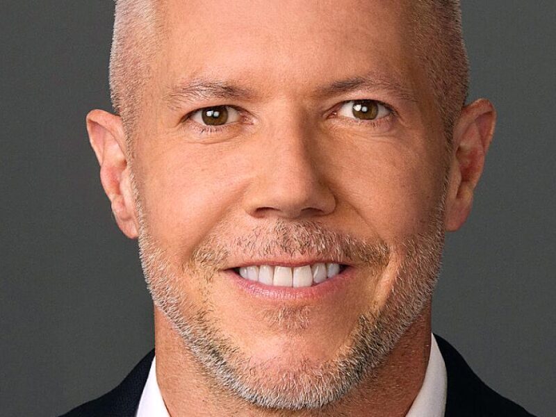 Portrait en gros plan de Paul Roeder, directeur de la communication de Disney, souriant, cheveux courts et clairs, barbe taillée, vêtu d'une veste sombre et d'une chemise blanche, sur un fond gris uni.