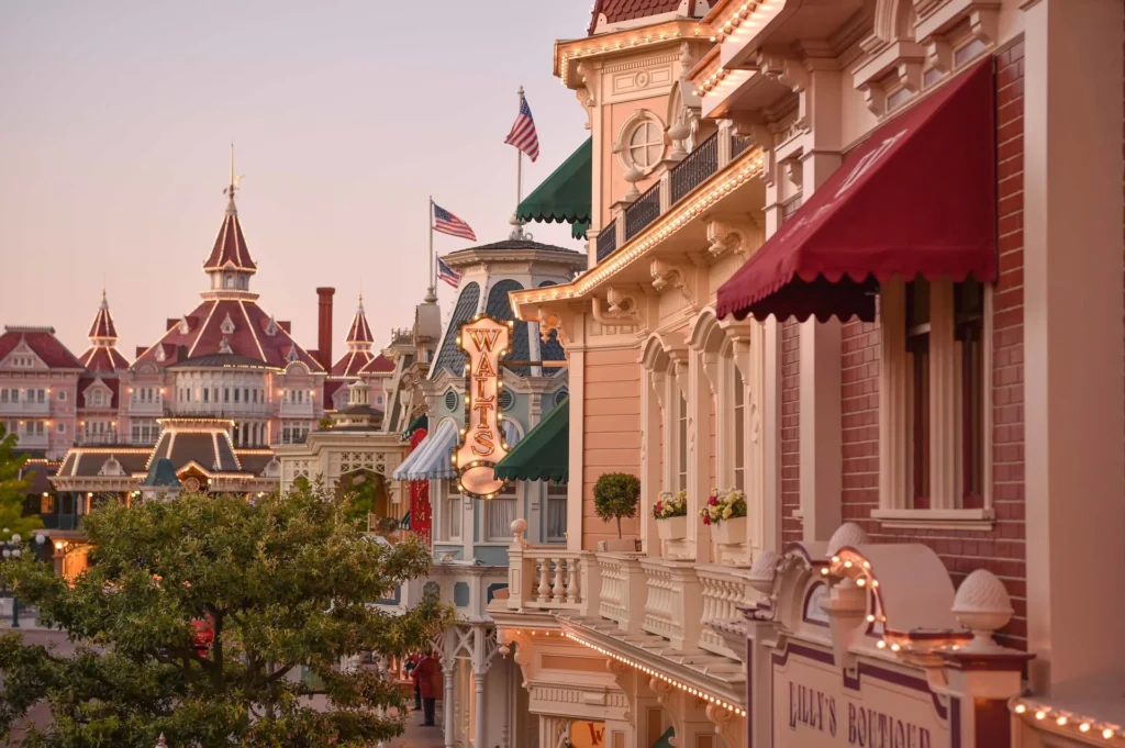 Une vue de Main Street U.S.A. à Disneyland Paris avec des bâtiments ornés, des drapeaux américains, des enseignes de magasins et des lumières décoratives au crépuscule - mettant en valeur la vision intemporelle du design d'Eddie Sotto.