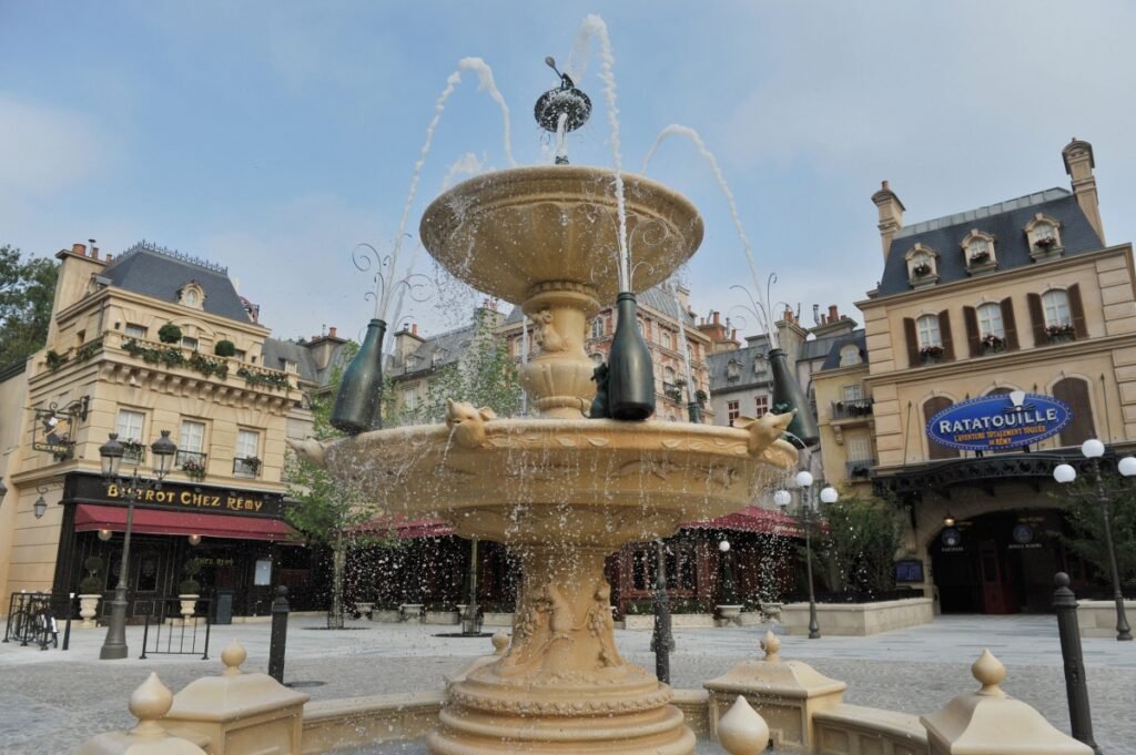 Une fontaine en pierre avec trois bouteilles décore une place entourée de bâtiments de style parisien, d'un bistrot et de la signalétique de l'attraction Ratatouille, capturant le charme et la culture locale des expériences Disneyland Paris.