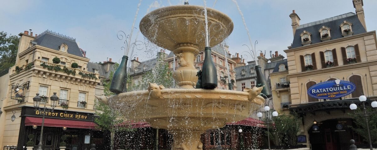 Une fontaine en pierre avec trois bouteilles décore une place entourée de bâtiments de style parisien, d'un bistrot et de la signalétique de l'attraction Ratatouille, capturant le charme et la culture locale des expériences Disneyland Paris.