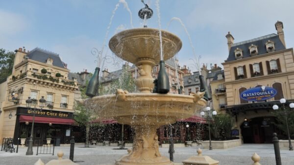 Une fontaine en pierre avec trois bouteilles décore une place entourée de bâtiments de style parisien, d'un bistrot et de la signalétique de l'attraction Ratatouille, capturant le charme et la culture locale des expériences Disneyland Paris.
