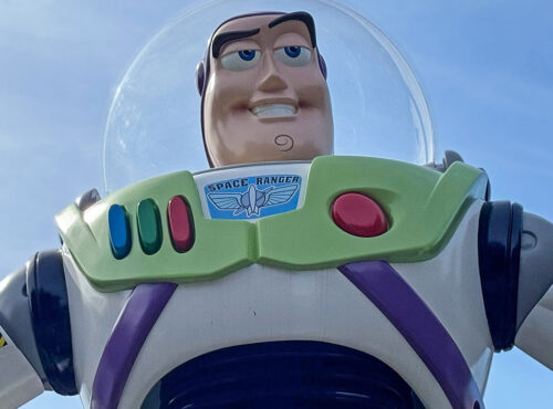 Gros plan d'une figurine de Buzz l'éclair dans sa combinaison spatiale blanche, verte et violette, sur fond de ciel bleu clair, rappelant l'attraction Buzz l'éclair laser de Disneyland Paris.
