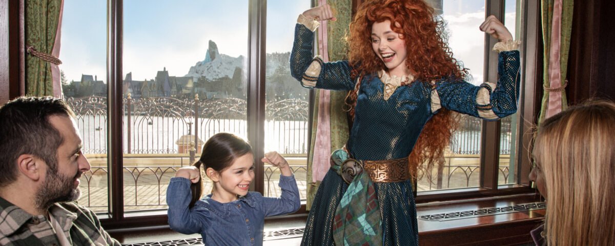 Une jeune fille et une femme en costume fléchissent les bras et sourient, tandis que deux adultes regardent depuis une table dans un restaurant à Disney Adventure World, avec de grandes fenêtres révélant un paysage de montagne, le 24 Juillet pendant la Cavalcade des Princesses Disney.