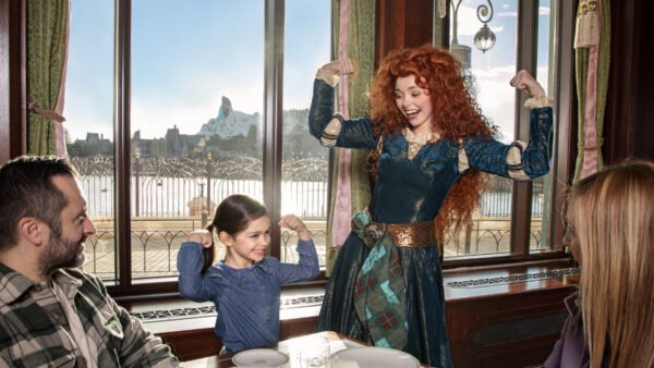 Une jeune fille et une femme en costume fléchissent les bras et sourient, tandis que deux adultes regardent depuis une table dans un restaurant à Disney Adventure World, avec de grandes fenêtres révélant un paysage de montagne, le 24 Juillet pendant la Cavalcade des Princesses Disney.