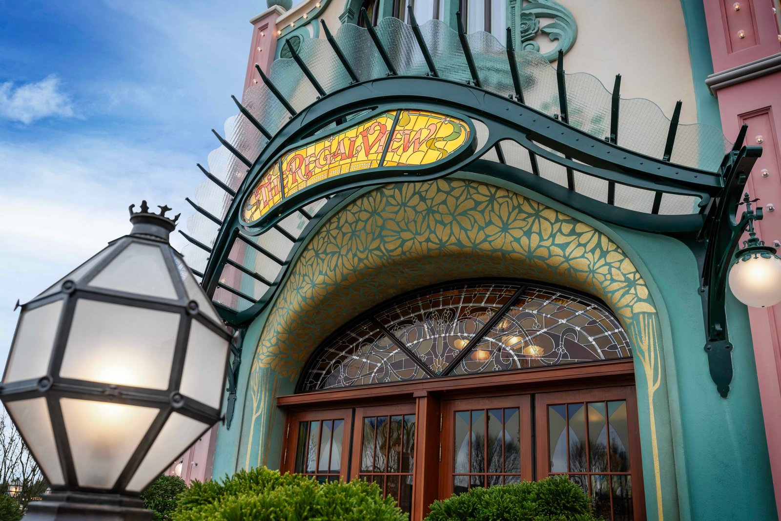 Entrée de style Art nouveau du restaurant Regal View, avec une enseigne en vitrail au-dessus de doubles portes en bois, des ornements réalisés par les Ateliers Schaffner et un lampadaire décoratif, évoquant le charme de Disney Adventure World.