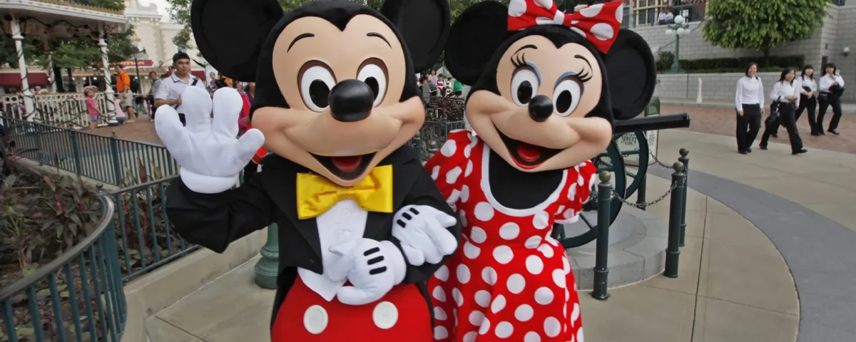 Les mascottes Mickey Mouse et Minnie Mouse posent et saluent sur une passerelle d'un parc à thème, avec en arrière-plan des personnes et des bâtiments reflétant les aménagements prévus par Disneyland pour l'accessibilité des personnes handicapées.