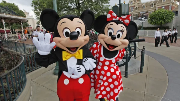 Les mascottes Mickey Mouse et Minnie Mouse posent et saluent sur une passerelle d'un parc à thème, avec en arrière-plan des personnes et des bâtiments reflétant les aménagements prévus par Disneyland pour l'accessibilité des personnes handicapées.