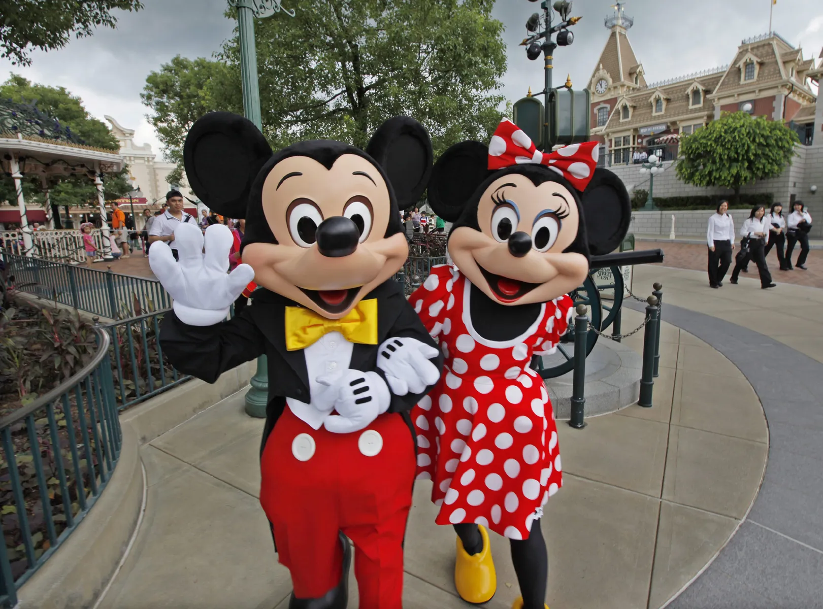 Les mascottes Mickey Mouse et Minnie Mouse posent et saluent sur une passerelle d'un parc à thème, avec en arrière-plan des personnes et des bâtiments reflétant les aménagements prévus par Disneyland pour l'accessibilité des personnes handicapées.