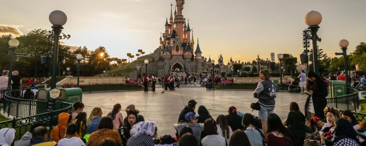 Une foule de personnes assises et debout devant un château de Disneyland Paris au coucher du soleil, avec des lampadaires et des clôtures décoratives visibles, soulignant l'atmosphère vibrante du parc et son rôle dans la création d'emplois.