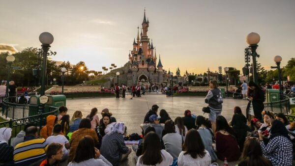 Une foule de personnes assises et debout devant un château de Disneyland Paris au coucher du soleil, avec des lampadaires et des clôtures décoratives visibles, soulignant l'atmosphère vibrante du parc et son rôle dans la création d'emplois.