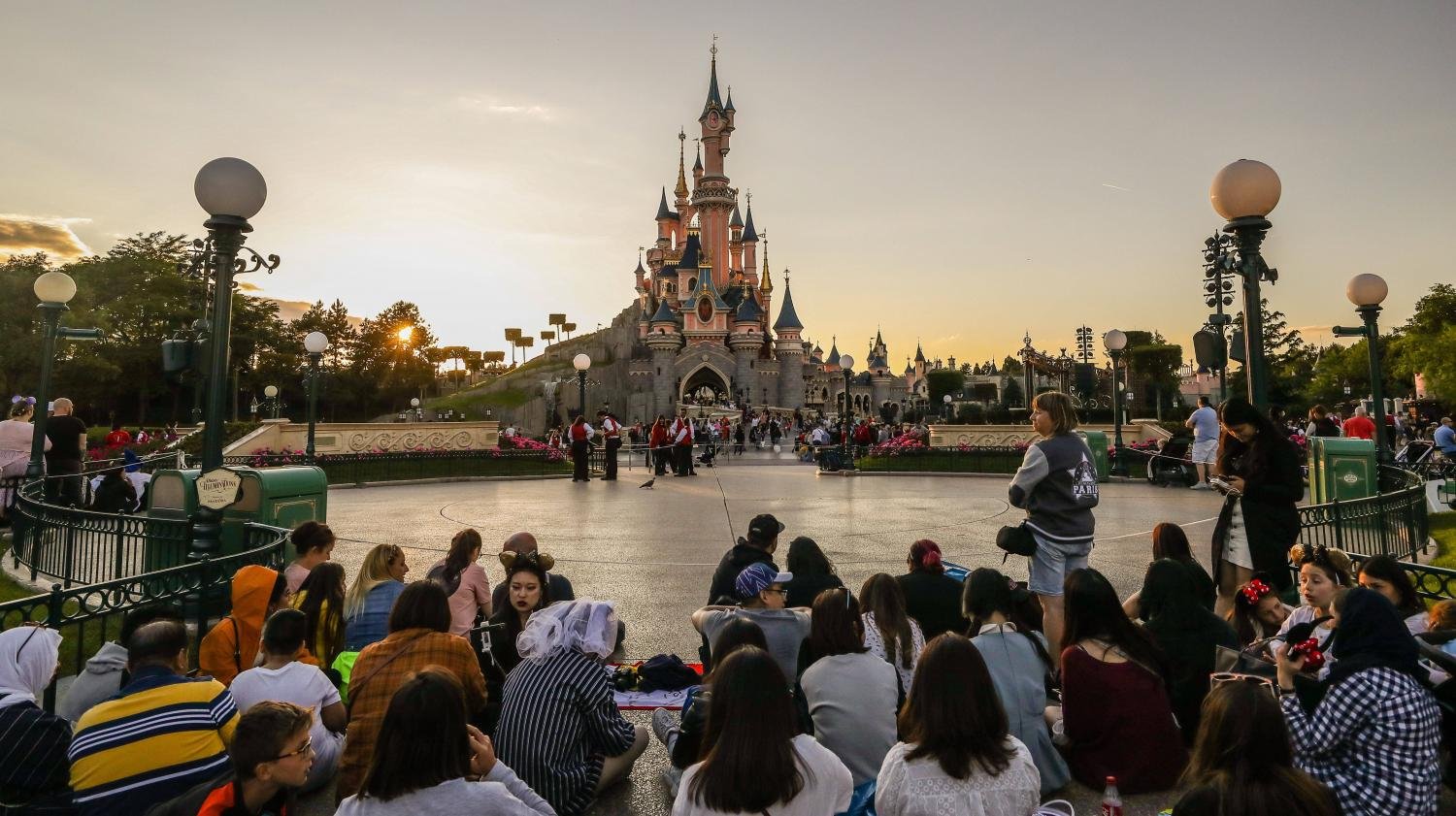 Une foule de personnes assises et debout devant un château de Disneyland Paris au coucher du soleil, avec des lampadaires et des clôtures décoratives visibles, soulignant l'atmosphère vibrante du parc et son rôle dans la création d'emplois.