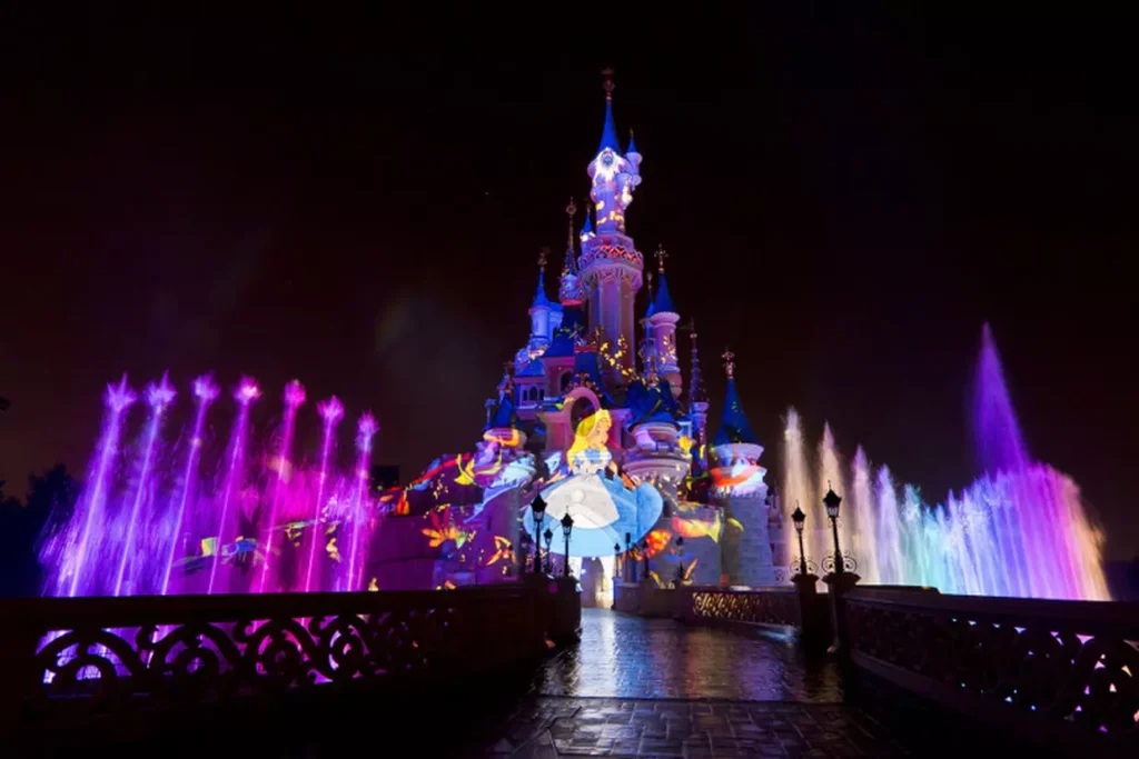 Le château de la Belle au bois dormant de Disneyland Paris s'illumine la nuit avec des lumières colorées, des fontaines d'eau et une image projetée d'Alice dans "Alice au pays des merveilles", créant une présence magique qui captive la perception des habitants et des visiteurs.
