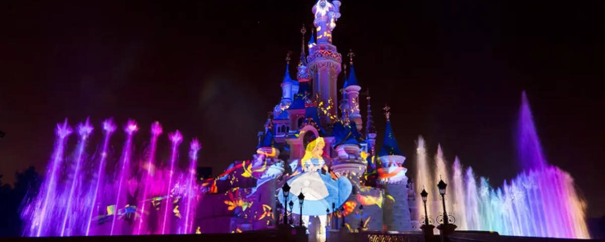Le château de la Belle au bois dormant de Disneyland Paris s'illumine la nuit avec des lumières colorées, des fontaines d'eau et une image projetée d'Alice dans "Alice au pays des merveilles", créant une présence magique qui captive la perception des habitants et des visiteurs.