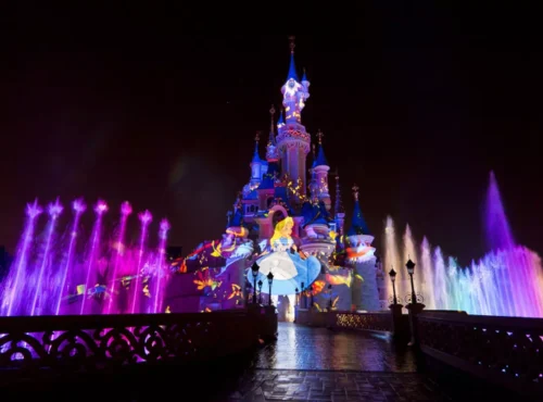 Le château de la Belle au bois dormant de Disneyland Paris s'illumine la nuit avec des lumières colorées, des fontaines d'eau et une image projetée d'Alice dans "Alice au pays des merveilles", créant une présence magique qui captive la perception des habitants et des visiteurs.