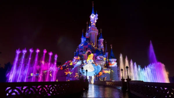 Le château de la Belle au bois dormant de Disneyland Paris s'illumine la nuit avec des lumières colorées, des fontaines d'eau et une image projetée d'Alice dans "Alice au pays des merveilles", créant une présence magique qui captive la perception des habitants et des visiteurs.