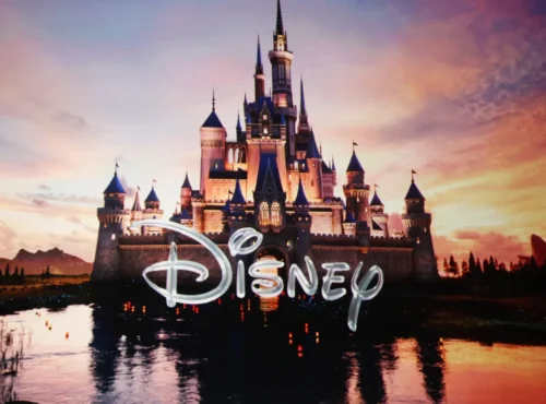 Un château aux flèches bleues se reflète dans un lac au coucher du soleil, tandis que le mot "Disney" apparaît en gros caractères sur l'image, soulignant subtilement la récente restructuration de l'entreprise.