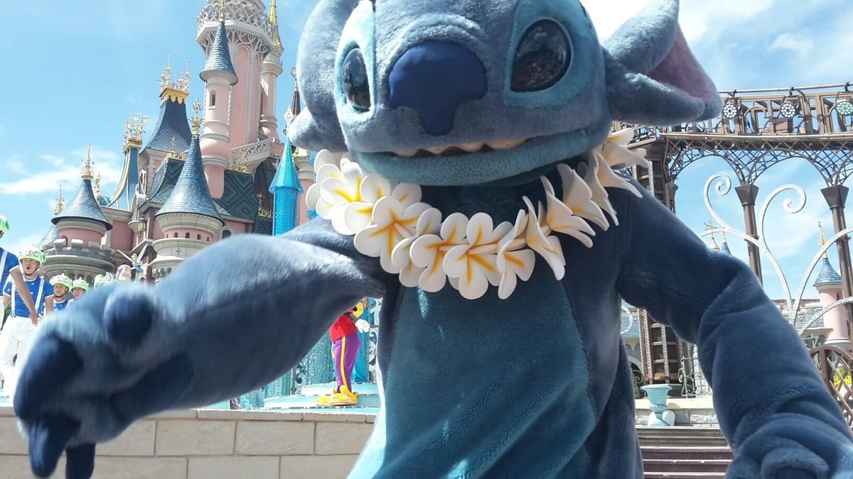 Une personne déguisée en Stitch et portant un lei de fleurs s'approche de l'appareil photo à Disneyland Paris, avec un château et des artistes en arrière-plan, à l'occasion des Stitch Days.