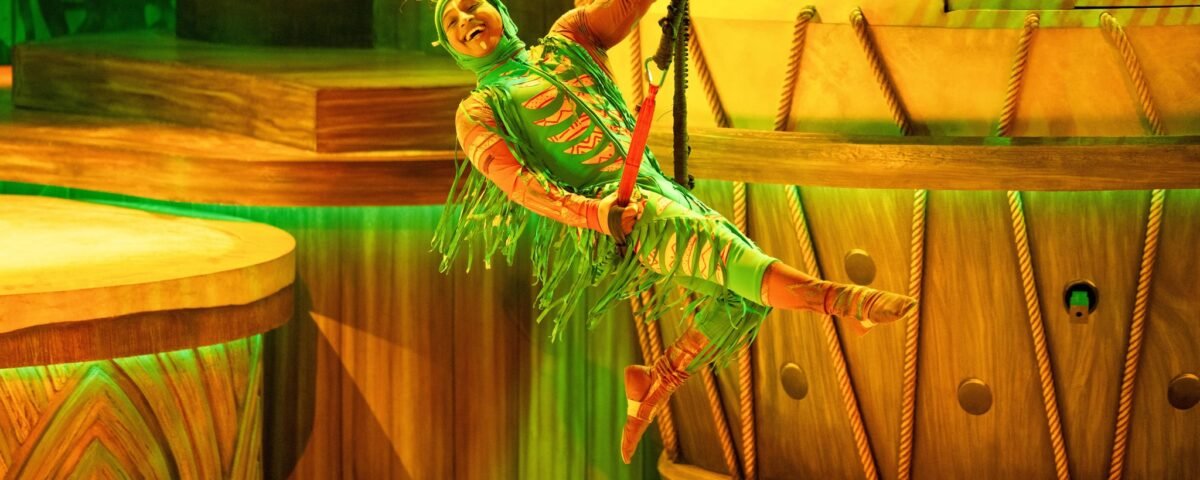 Un artiste vêtu d'un costume coloré se balance sur une corde pendant le spectacle Le Roi Lion à Disneyland Paris, éblouissant le public au milieu de grands décors en bois et d'un éclairage vert vibrant.