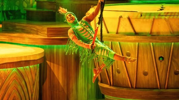 Un artiste vêtu d'un costume coloré se balance sur une corde pendant le spectacle Le Roi Lion à Disneyland Paris, éblouissant le public au milieu de grands décors en bois et d'un éclairage vert vibrant.