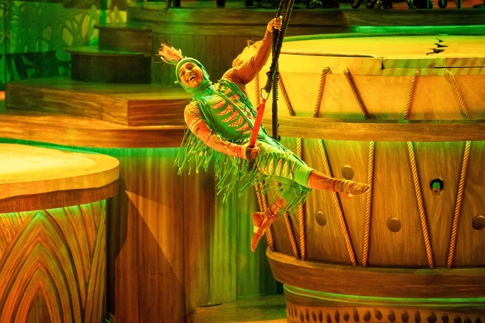 Un artiste vêtu d'un costume coloré se balance sur une corde pendant le spectacle Le Roi Lion à Disneyland Paris, éblouissant le public au milieu de grands décors en bois et d'un éclairage vert vibrant.