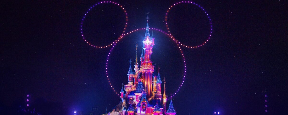 Un château illuminé de nuit avec des lumières colorées et des drones formant une grande tête de Mickey Mouse dans le ciel, mettant en valeur la magie de Disneyland Paris en collaboration avec d'autres parcs Disney.