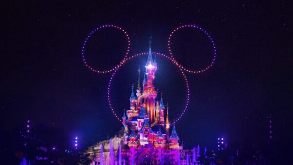 Un château illuminé de nuit avec des lumières colorées et des drones formant une grande tête de Mickey Mouse dans le ciel, mettant en valeur la magie de Disneyland Paris en collaboration avec d'autres parcs Disney.