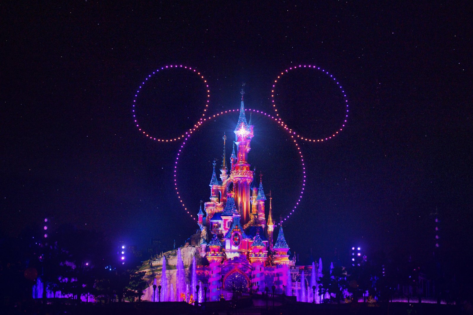 Un château illuminé de nuit avec des lumières colorées et des drones formant une grande tête de Mickey Mouse dans le ciel, mettant en valeur la magie de Disneyland Paris en collaboration avec d'autres parcs Disney.
