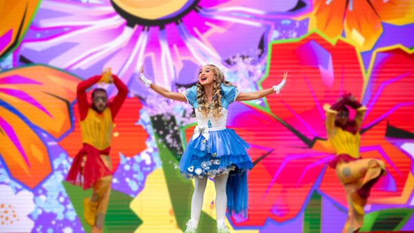 Une artiste vêtue d'une robe bleue et blanche chante sur scène devant des accessoires floraux colorés et surdimensionnés, avec deux danseuses en costumes vibrants derrière elle, lors du spectacle Alice Et La Reine De Cœur de Disney Adventure World : Retour Au Pays Des Merveilles.