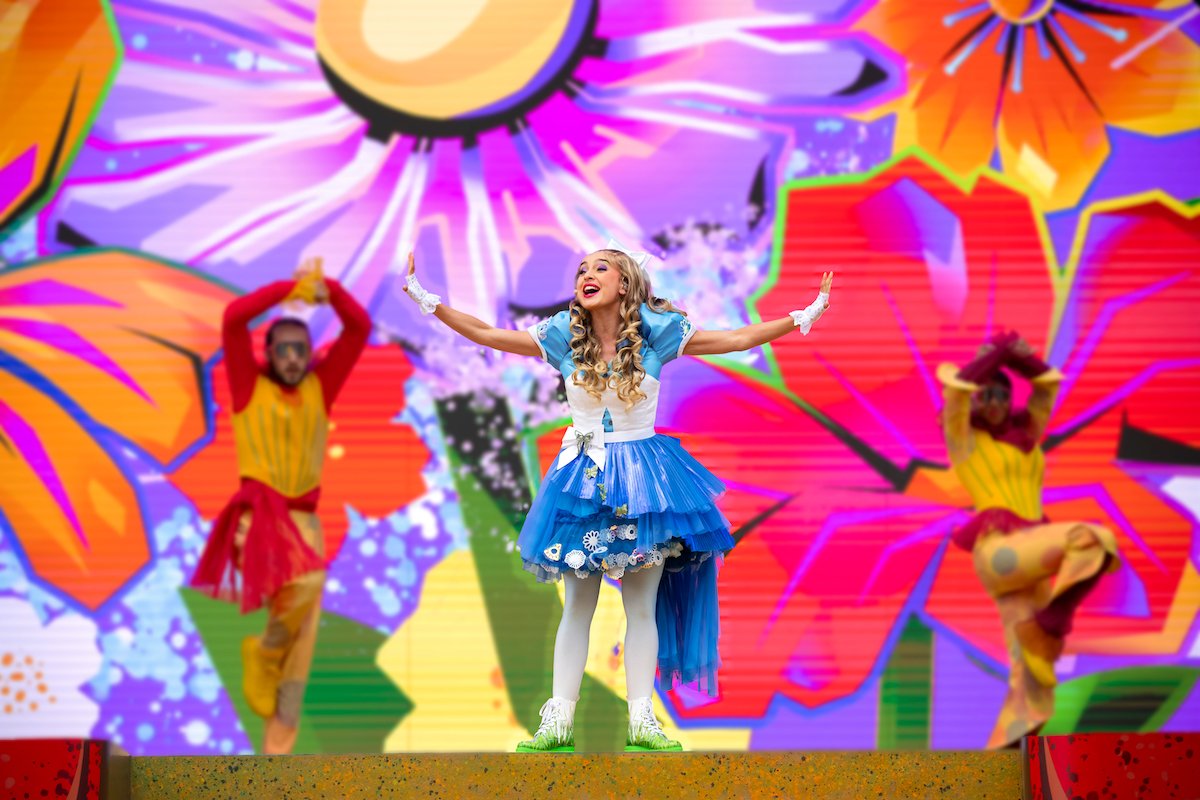 Une artiste vêtue d'une robe bleue et blanche chante sur scène devant des accessoires floraux colorés et surdimensionnés, avec deux danseuses en costumes vibrants derrière elle, lors du spectacle Alice Et La Reine De Cœur de Disney Adventure World : Retour Au Pays Des Merveilles.